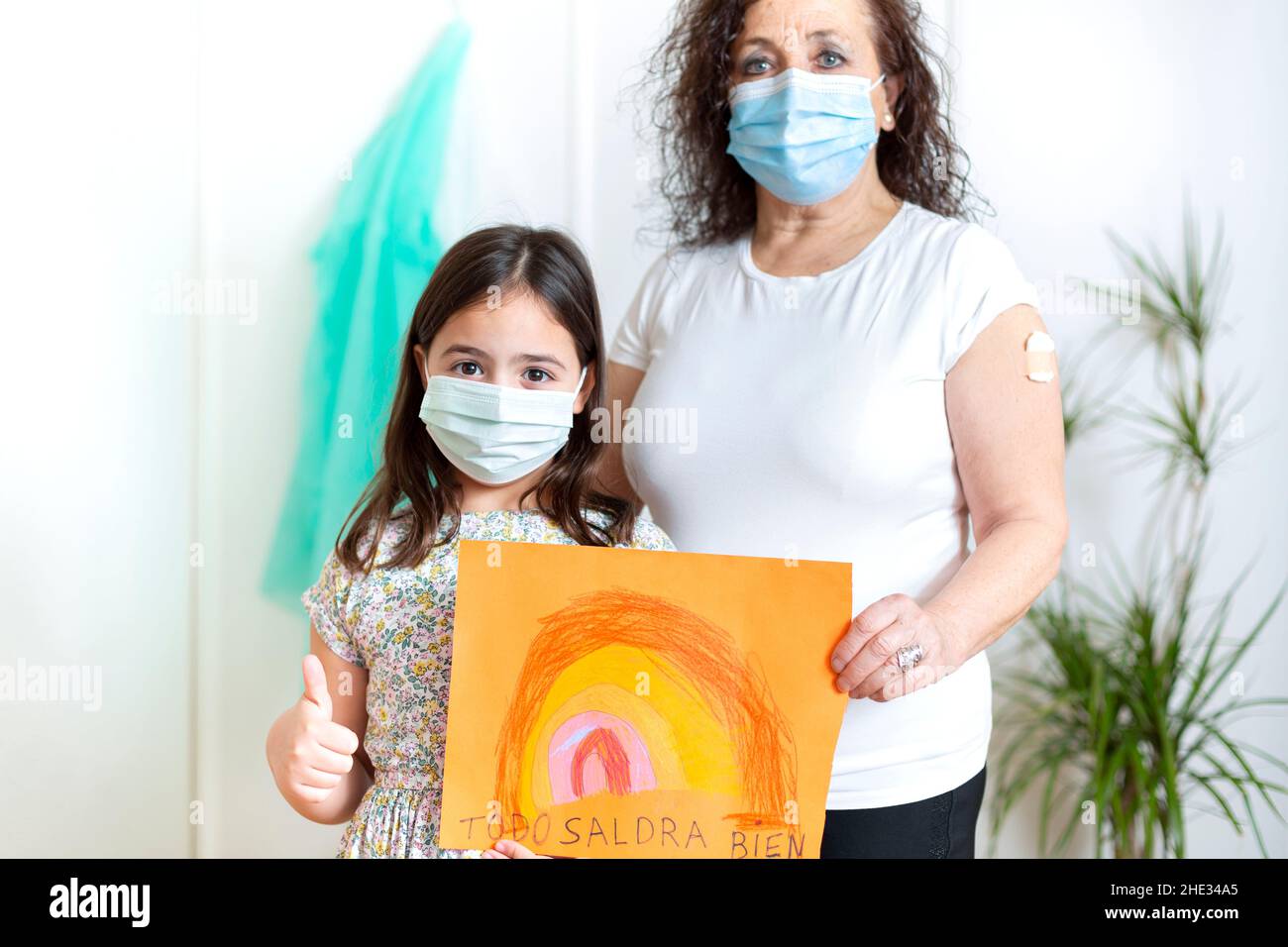 Niña y mujer mayor, ambas con máscaras faciales, posando con un cartel de arco iris dibujado a mano con el eslogan 'Todo estará bien'. Foto de stock