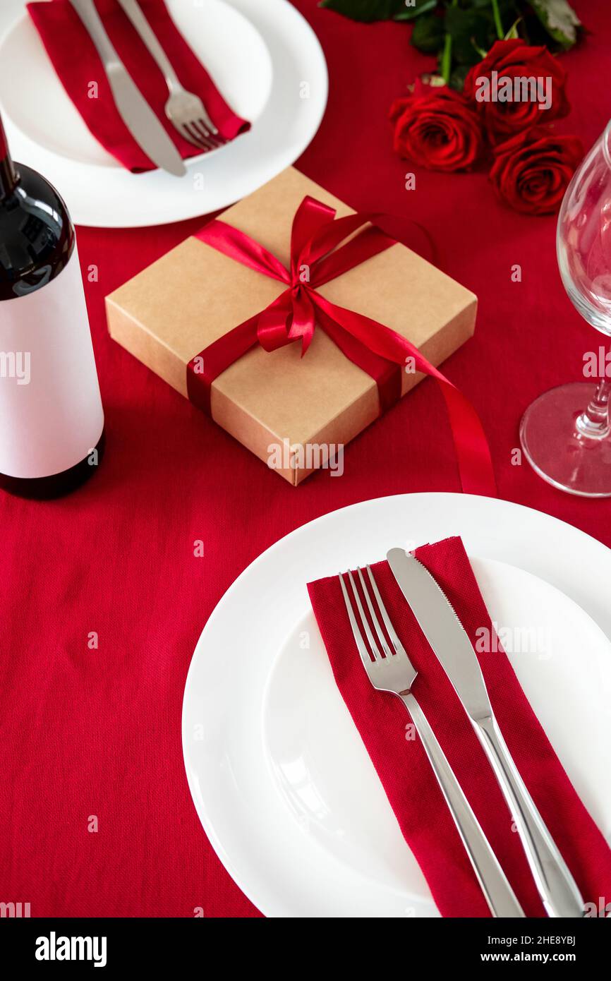 Cena romántica de San Valentín para dos mesas con vistas a la cima. Mesa  con copa de vino, caja de regalo, vajilla, tenedor, cuchillo, flores de  color rosa sobre fondo rojo Fotografía