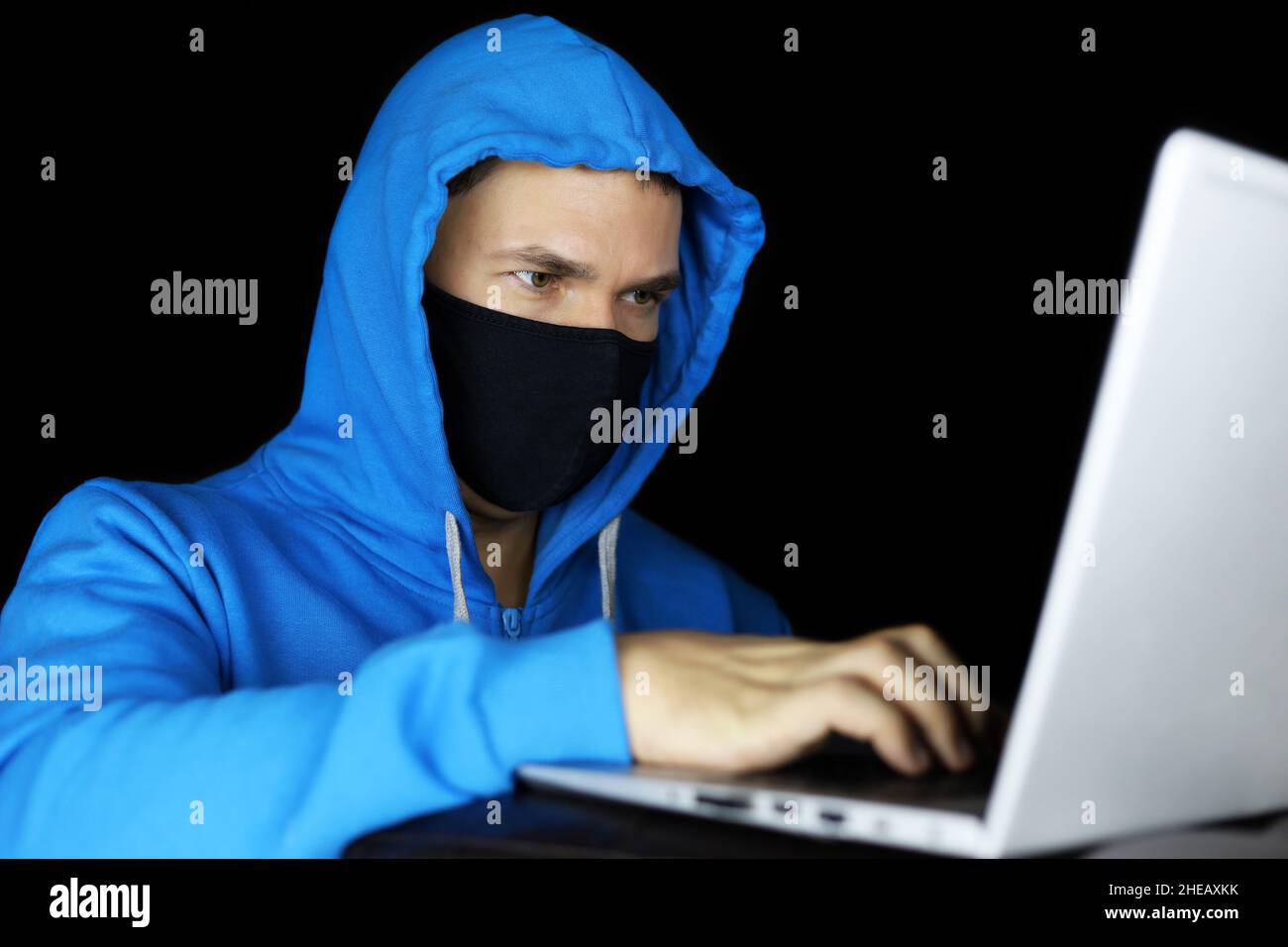 Hombre con máscara y sudadera con capucha azul sentado con un ordenador portátil sobre fondo negro. Concepto de delito cibernético, piratería informática y tecnología Foto de stock