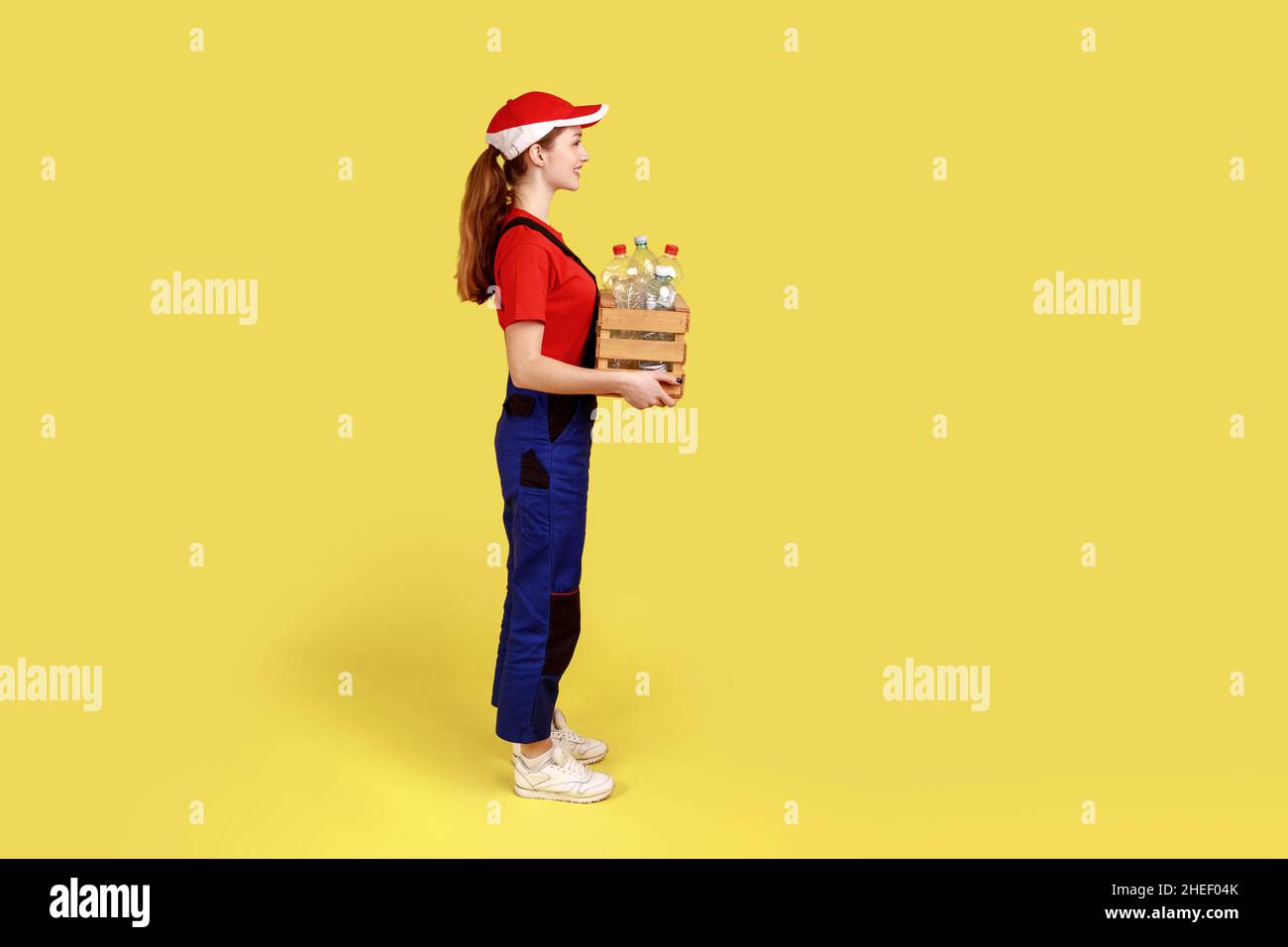 Vista lateral retrato de una voluntaria positiva satisfecha de pie con una caja llena de botellas de plástico vacías, con monos y gorra roja. Estudio en interior grabado aislado sobre fondo amarillo. Foto de stock
