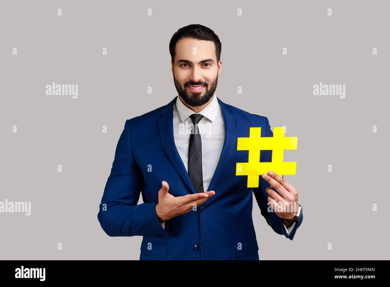 Retrato de sonriente hombre barbudo presentando amarillo hashtag, marcado de tendencias de blog, tema viral en la red social, con traje de estilo oficial. Estudio en interior grabado aislado sobre fondo gris. Foto de stock
