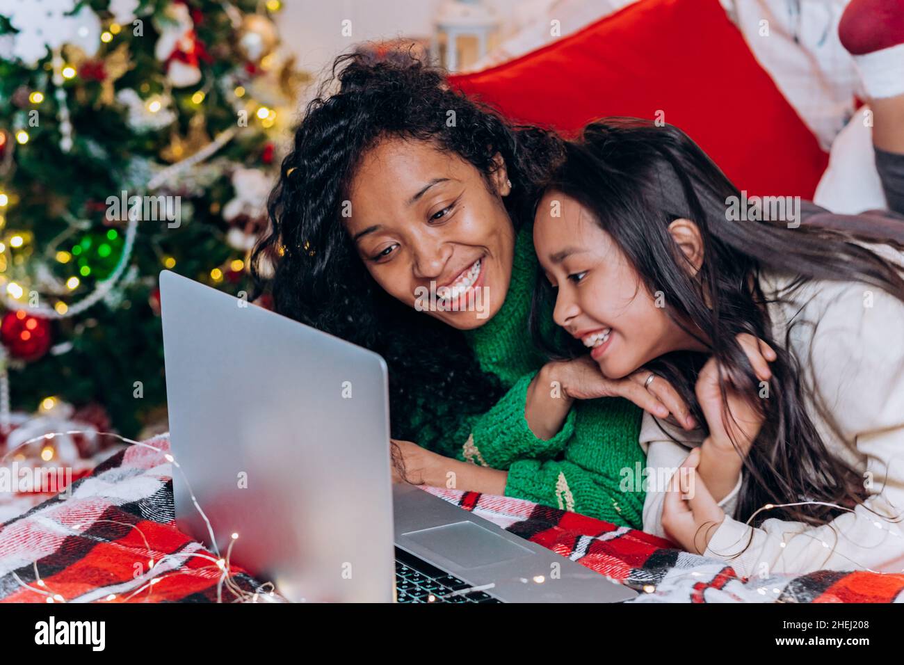 La madre negra de pelo rizado y la hija morena yacen en la cama cerca de decoraciones navideñas y se comunican mediante videollamada con el ordenador portátil Foto de stock