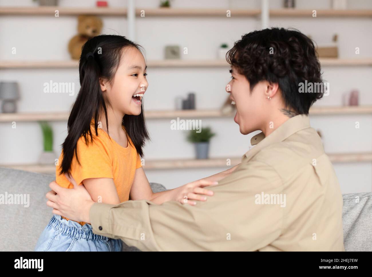 Feliz asiático millennial hombre y hija adolescente mirar el uno al otro con bocas abiertas, divertirse Foto de stock
