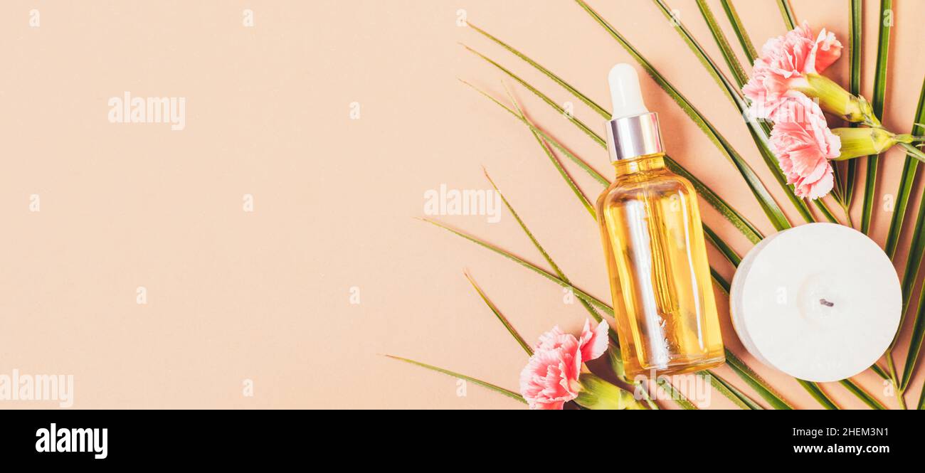 Botella de suero cosmético junto a flores frescas, vela y hoja de palma sobre fondo marrón con espacio de copia. Plantilla de banner de ancho largo Foto de stock