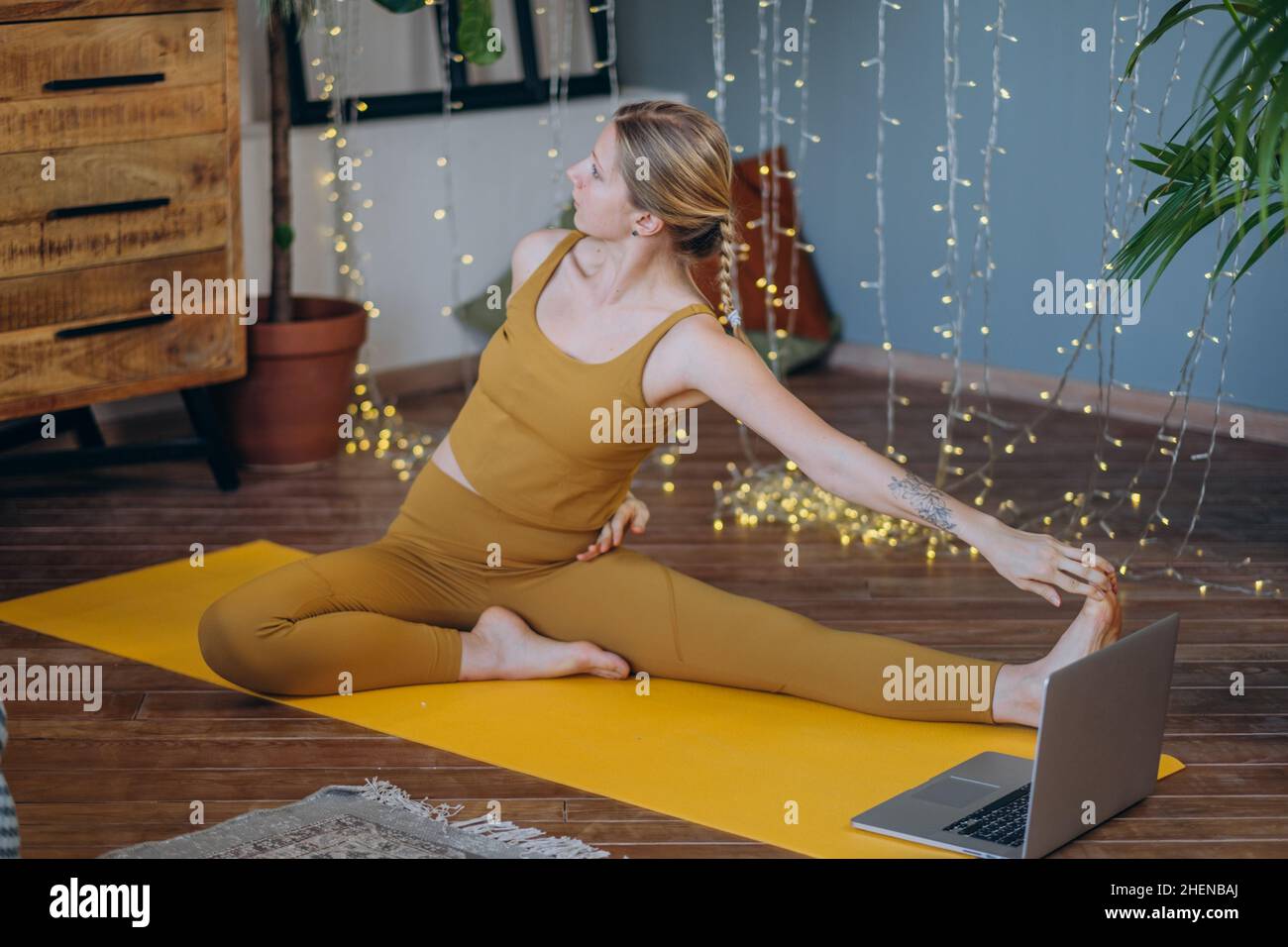Mujer joven en la parte superior y leggings hace ejercicios de estiramiento en la colchoneta de yoga ver tutorial en el portátil contra iluminar la guirnalda Foto de stock
