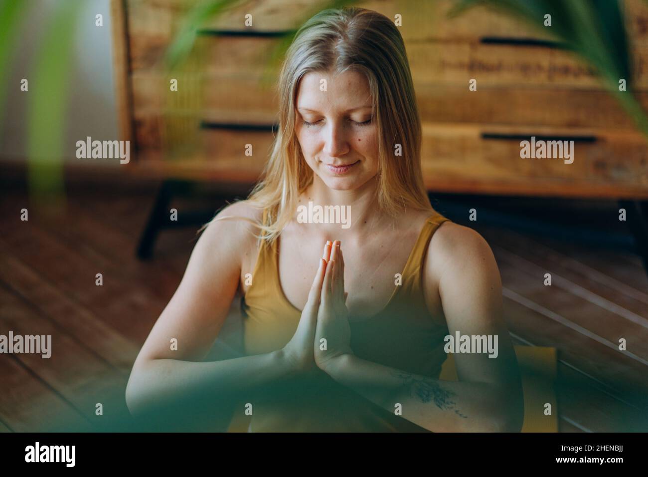 Mujer atlética con pelo rubio suelto y tatuajes en la parte superior y leggings medita con amplia sonrisa sentada en posición de loto en la colchoneta de yoga cerca de la vista Foto de stock