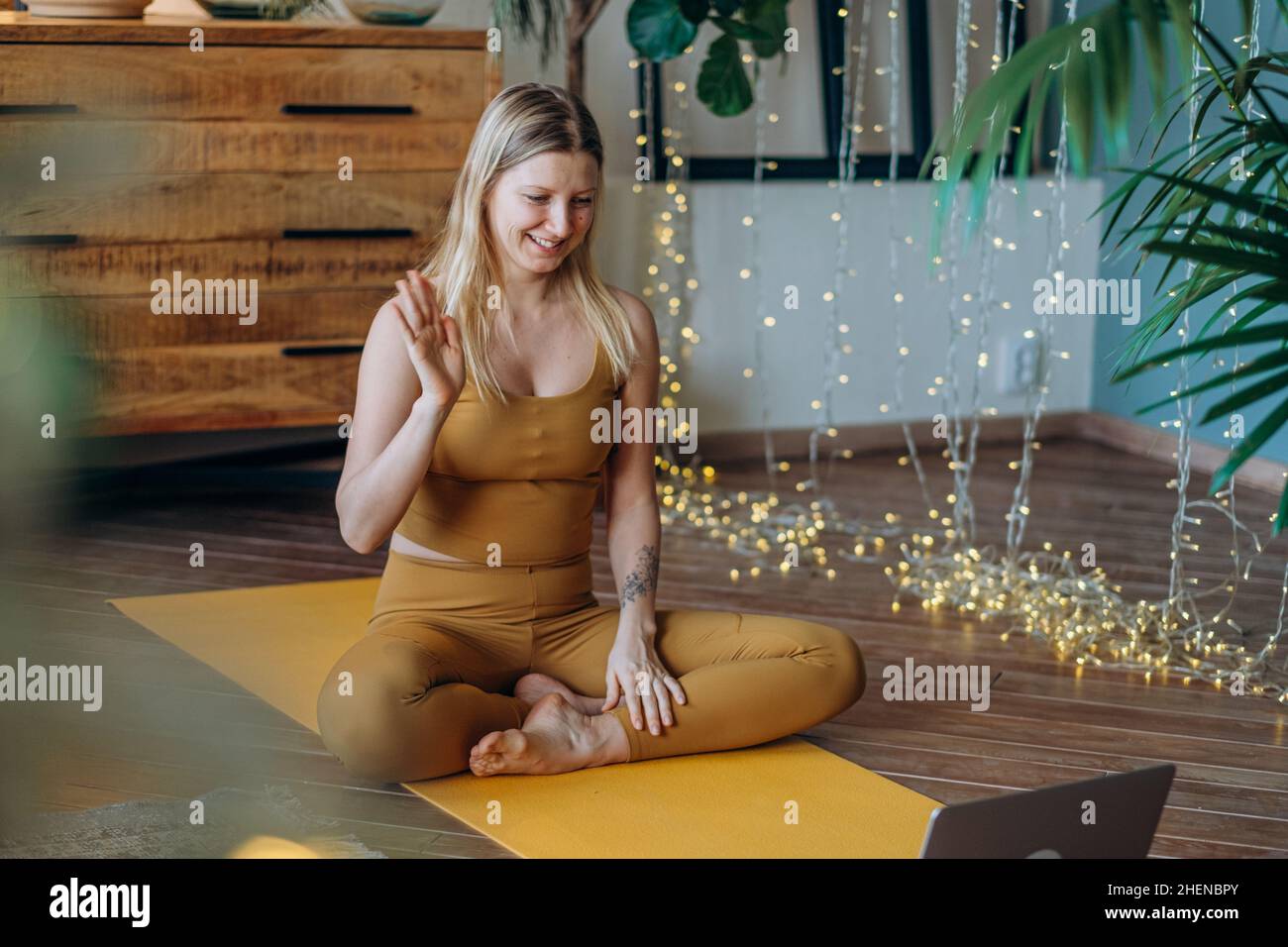 Mujer joven en la parte superior y leggings hace ejercicios de estiramiento en la colchoneta de yoga ver tutorial en el portátil contra iluminar la guirnalda Foto de stock