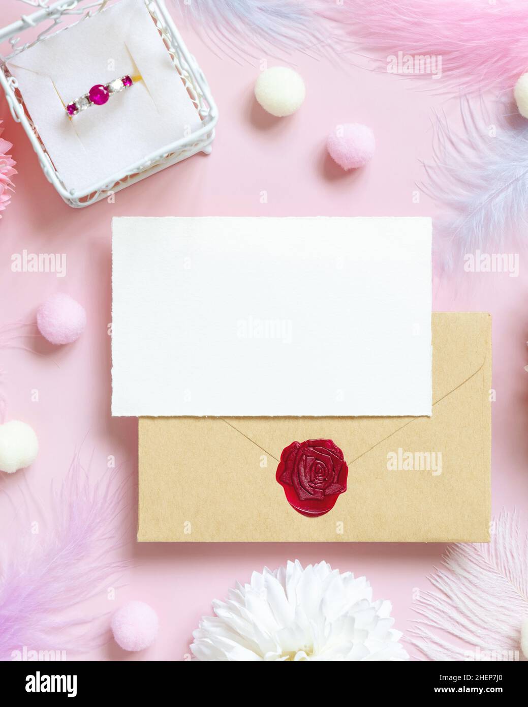 Tarjeta y sobre cerca de flores pastel, pompones, plumas y anillo en una  caja de regalo en la vista superior rosa. Escena romántica con sobre en  blanco y lugar para te Fotografía