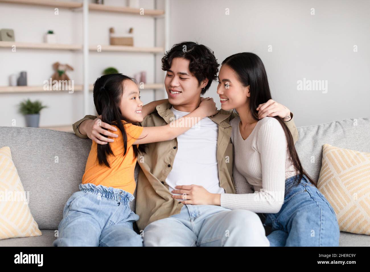 Positivo bastante joven chica japonesa, mamá y papá abrazando en el sofá divertirse, disfrutar de momento tierno Foto de stock