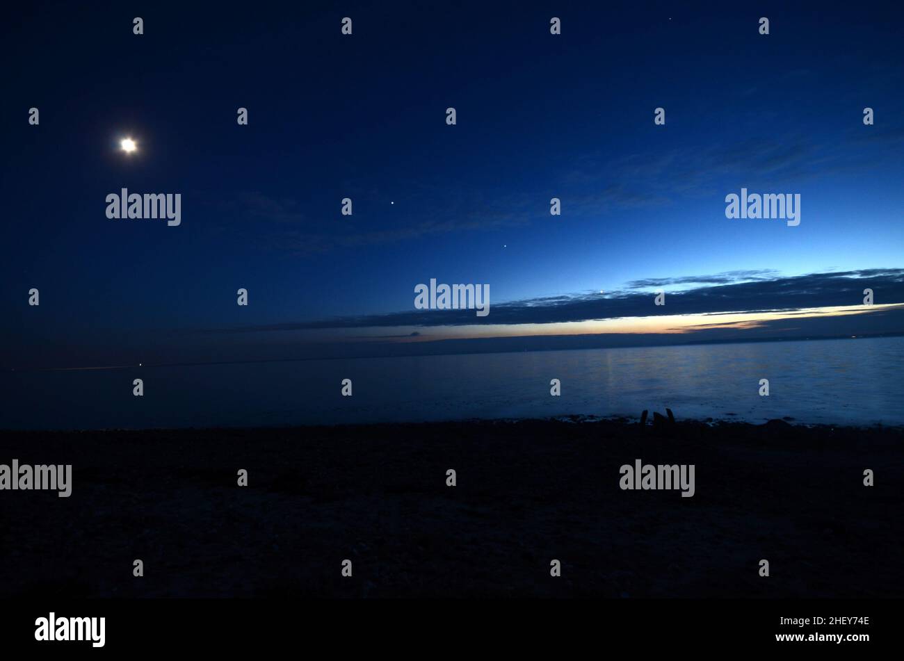 Vista sobre el mar al atardecer con la Luna, Júpiter, Saturno y Venus justo sobre las nubes. Los planetas parecen estar en una línea, esto se llama los ecli Foto de stock