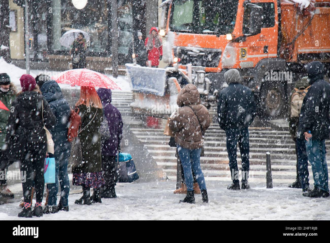 Belgrado, Serbia - 11 de enero de 2022: Personas esperando en el cruce de carreteras en una calle nevada de la ciudad durante una fuerte caída de nieve, y un camión de remoción de nieve está conduciendo pa Foto de stock