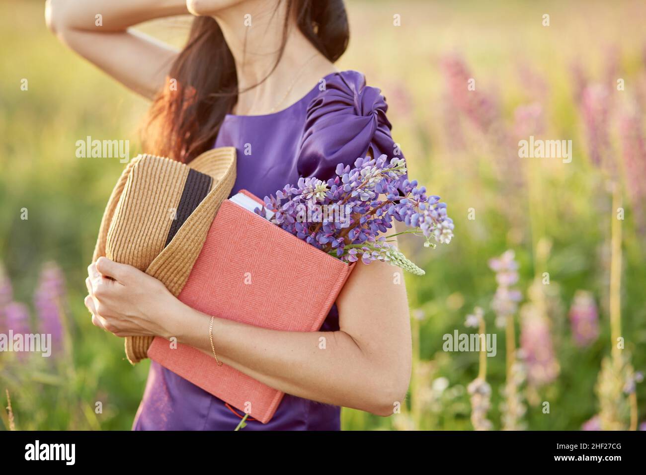 Mujer en el campo de verano con ramo de flores silvestres - altramuces, sombrero y cuaderno rosa. Proximidad a la naturaleza, concepto de auto-descubrimiento. Concepto de relax y bienestar. Día soleado, concepto de buen humor. Foto de stock