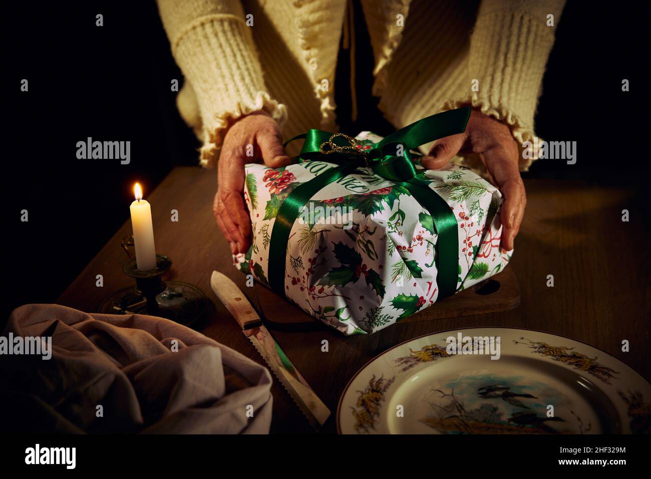 Una mujer sostiene con sus manos una torta de Navidad en una caja de regalo acostada en una tabla en una mesa de madera cerca de un cuchillo, una servilleta, un plato y una vela ardiente. Foto de stock