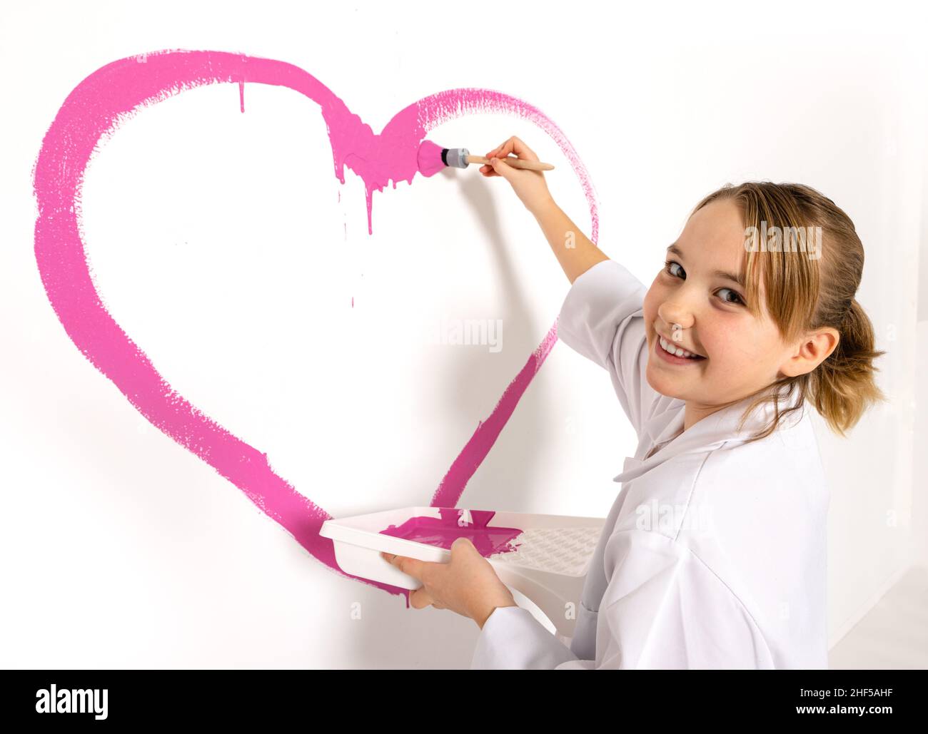 Niña de 10 años pintando un corazón rosa con un cepillo en la pared. La chica está mirando a la cámara con una sonrisa feliz. Foto de stock