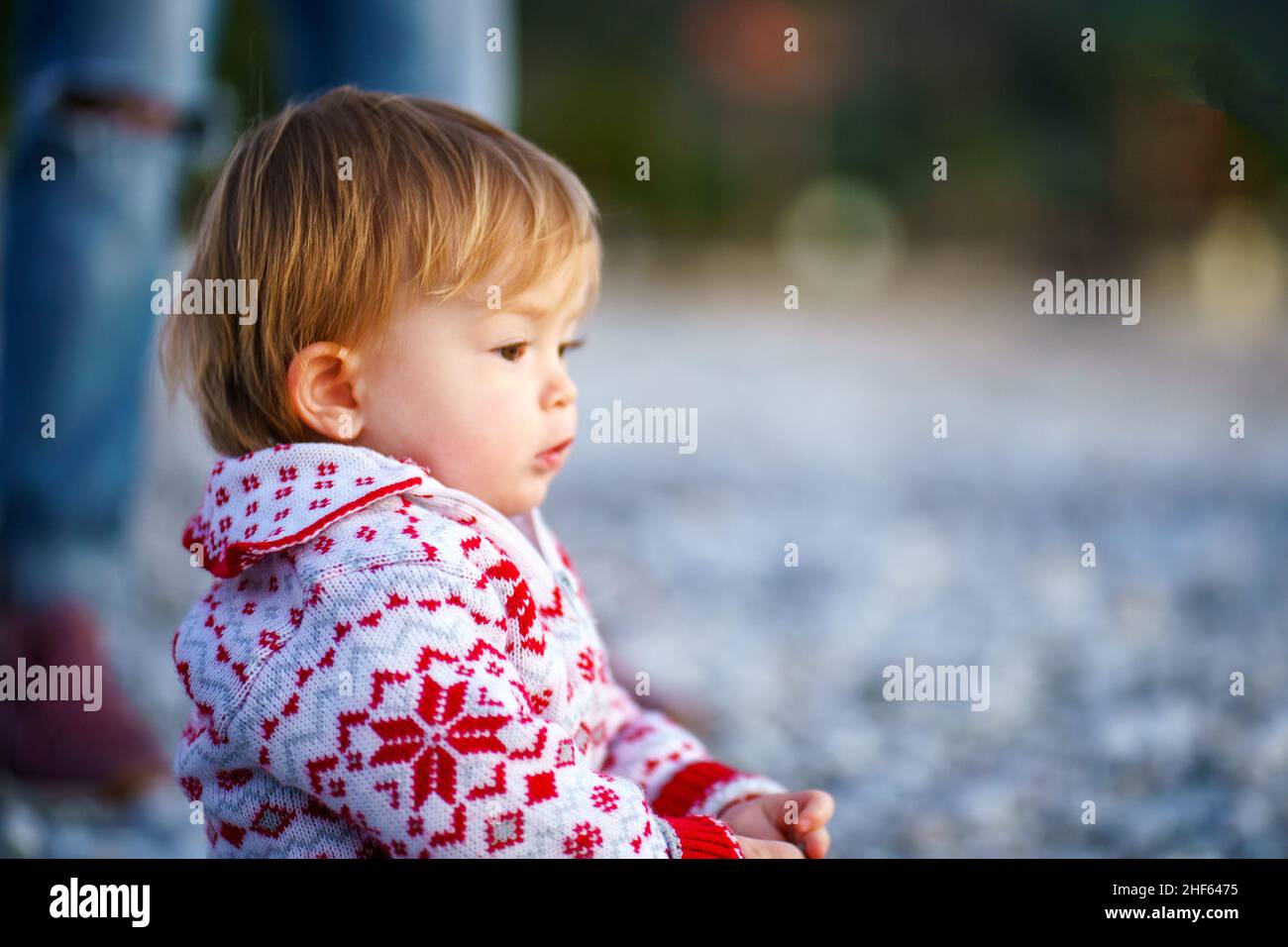 Un lindo bebé en un hermoso suéter en la orilla del mar de invierno. Hermoso niño en la naturaleza. El niño se sienta en perfil en una playa de piedra. Foto de stock
