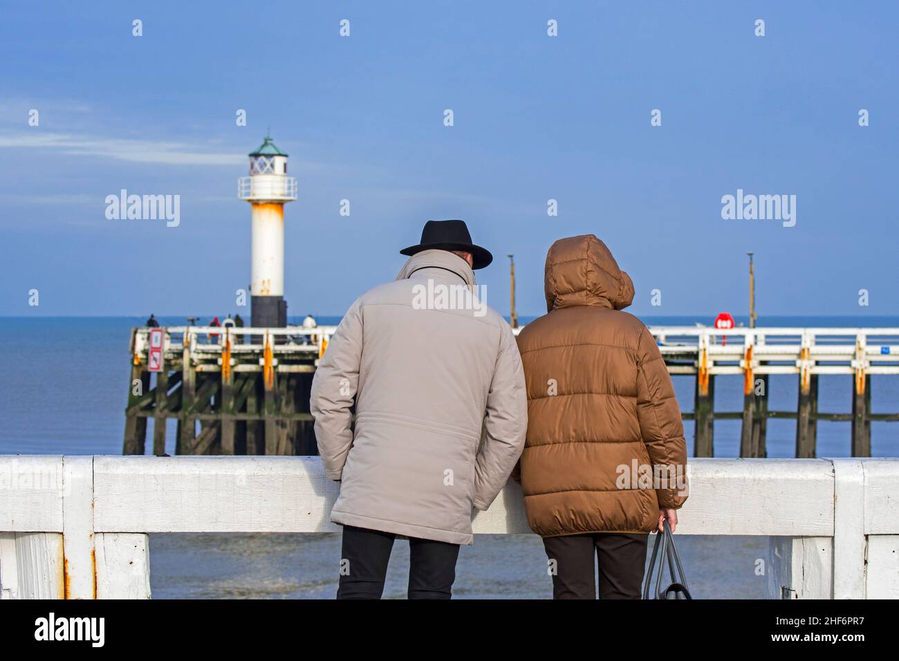 Pareja de ancianos vestida con abrigos en el muelle de madera en la entrada del puerto Nieuwpoort / Nieuport a lo largo de la costa belga del Mar del Norte en invierno, Wes Foto de stock