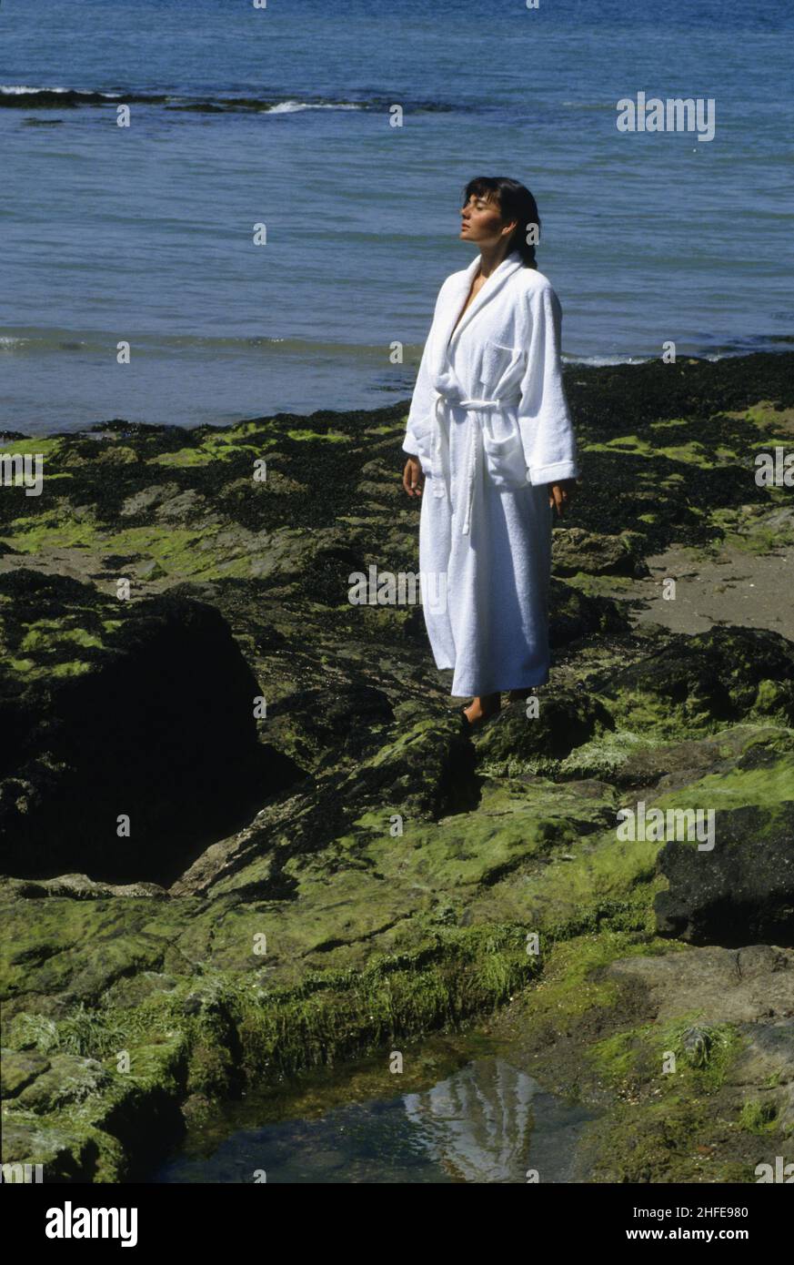 mujer joven de pelo oscuro ponerse de pie con bata de talasoterapia blanca caminando sobre la roca de la playa un montón de algas marinas Foto de stock