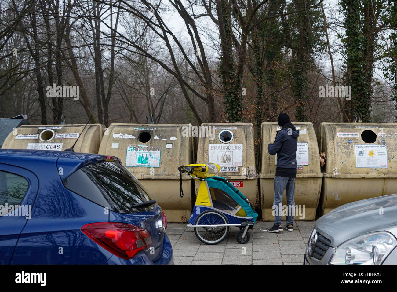 Un joven ha traído vacíos a los contenedores de reciclaje de su vecindario con un carro para bebés y les lanza botellas vacías. Foto de stock
