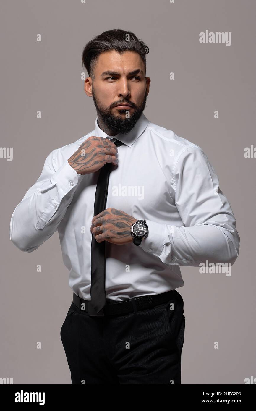 Hombre obado hispano con las manos tatuadas en camisa blanca y pantalón  negro corbata de ajuste y mirar hacia fuera mientras que de pie contra el  respaldo gris Fotografía de stock -