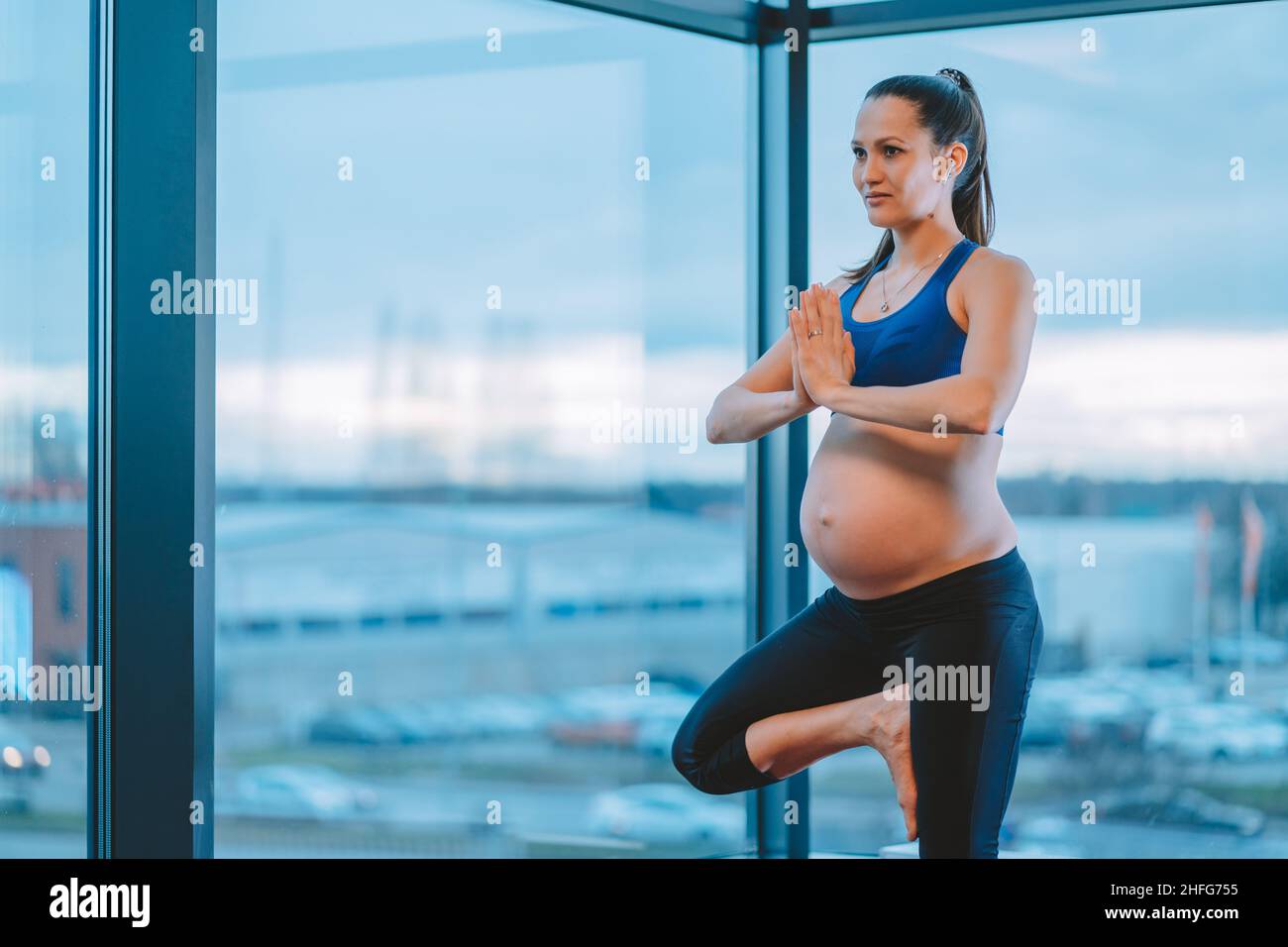 Mujer embarazada con las manos en gesto namaste de pie en la postura de árbol y practicar yoga en habitación con paredes de vidrio Foto de stock