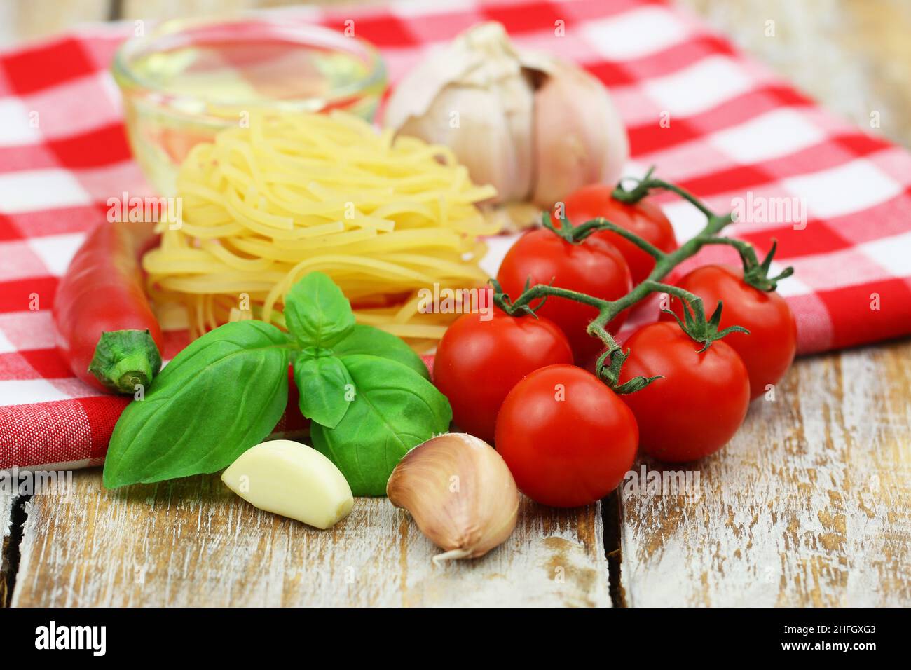 Ingredientes para cocinar pasta: Tagliatelle, tomates cherry, ajo, albahaca fresca, chile y un recipiente de aceite de oliva sobre un paño a cuadros Foto de stock