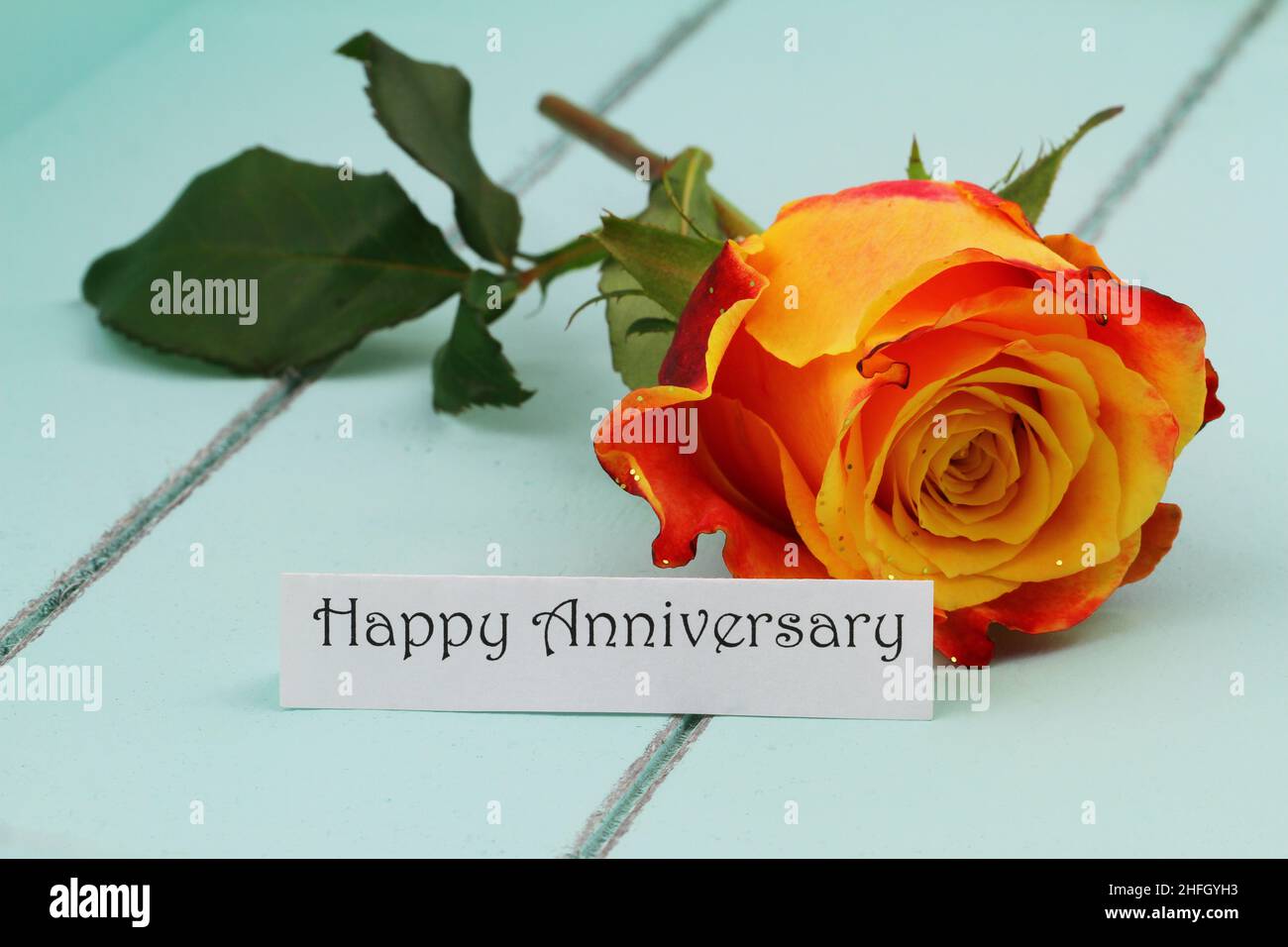 Tarjeta de feliz aniversario con una rosa roja y amarilla salpicada de brillos sobre una superficie de madera azul Foto de stock