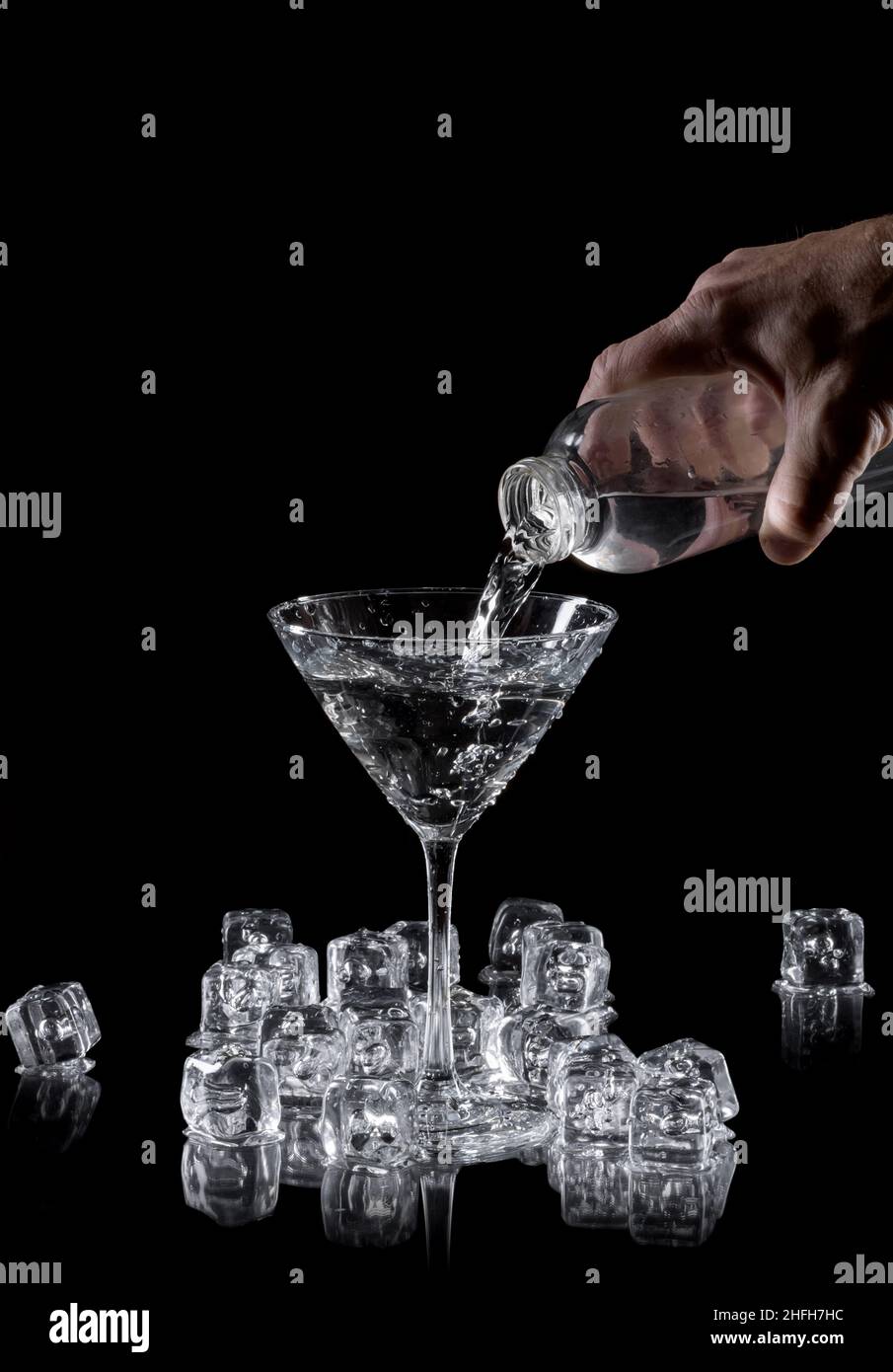 la mano de un hombre vertiendo ginebra o agua en un vaso de martini rodeado de cubitos de hielo sobre un fondo negro reflectante, con espacio de copia Foto de stock