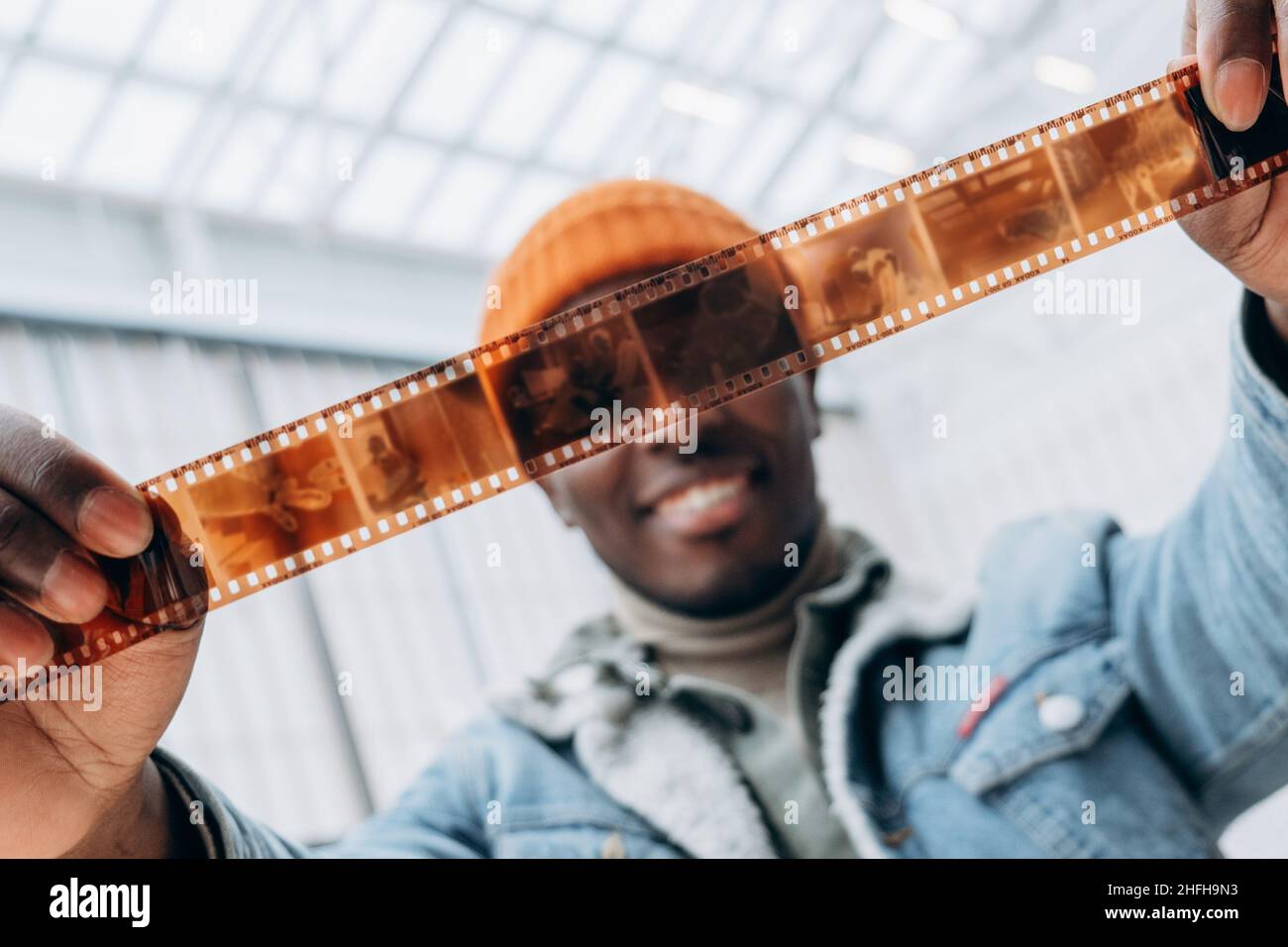 Emotivo feliz hombre afroamericano fotógrafo con chaqueta vaquera mira la película vintage en la sala de luz brillante extrema primer plano Foto de stock