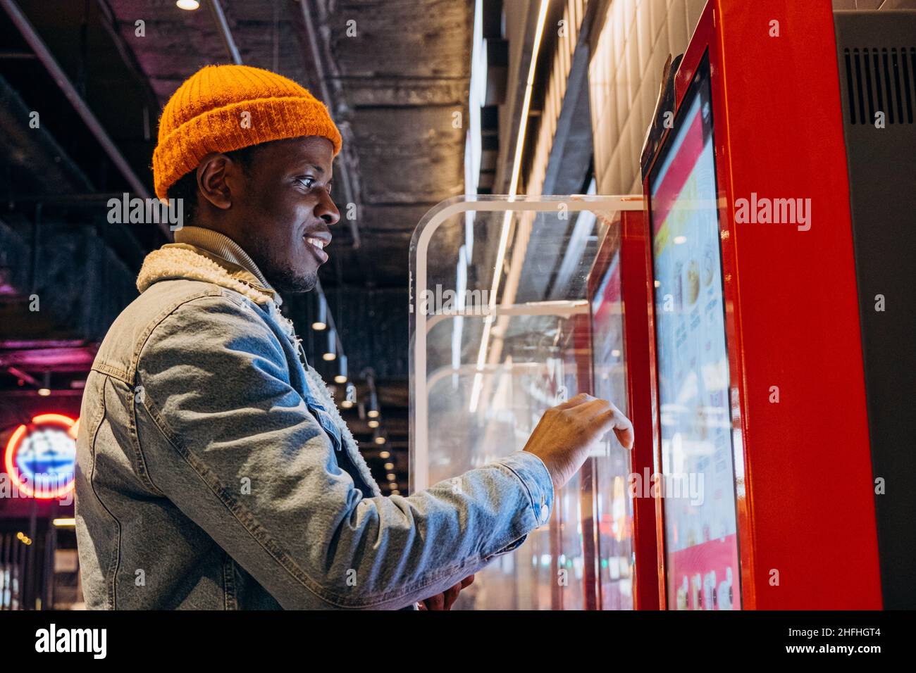 Un tipo afroamericano positivo con gorro de punto naranja con auriculares inalámbricos pide comida a través de un quiosco de autoservicio en el café Foto de stock