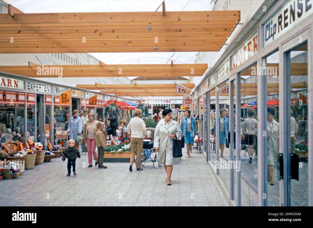 Compras encubiertas en el Tarup Center en las afueras de Odense, Funen, Dinamarca, en 1970. Las señales rojas y blancas indican que es tiempo de venta de verano (sommer udsalg). El uso de madera en el diseño del centro es evidente. El centro abrió en 1968 con 21 tiendas y fue el primer centro comercial cubierto en Funen. Ahora se ha ampliado mucho con más del doble del número de tiendas en el sitio y ha sido ampliamente remodelado. Odense es la ciudad principal de la isla de Funen. Esta imagen proviene de una transparencia de color amateur de 35mm – una fotografía vintage de 1970s. Foto de stock