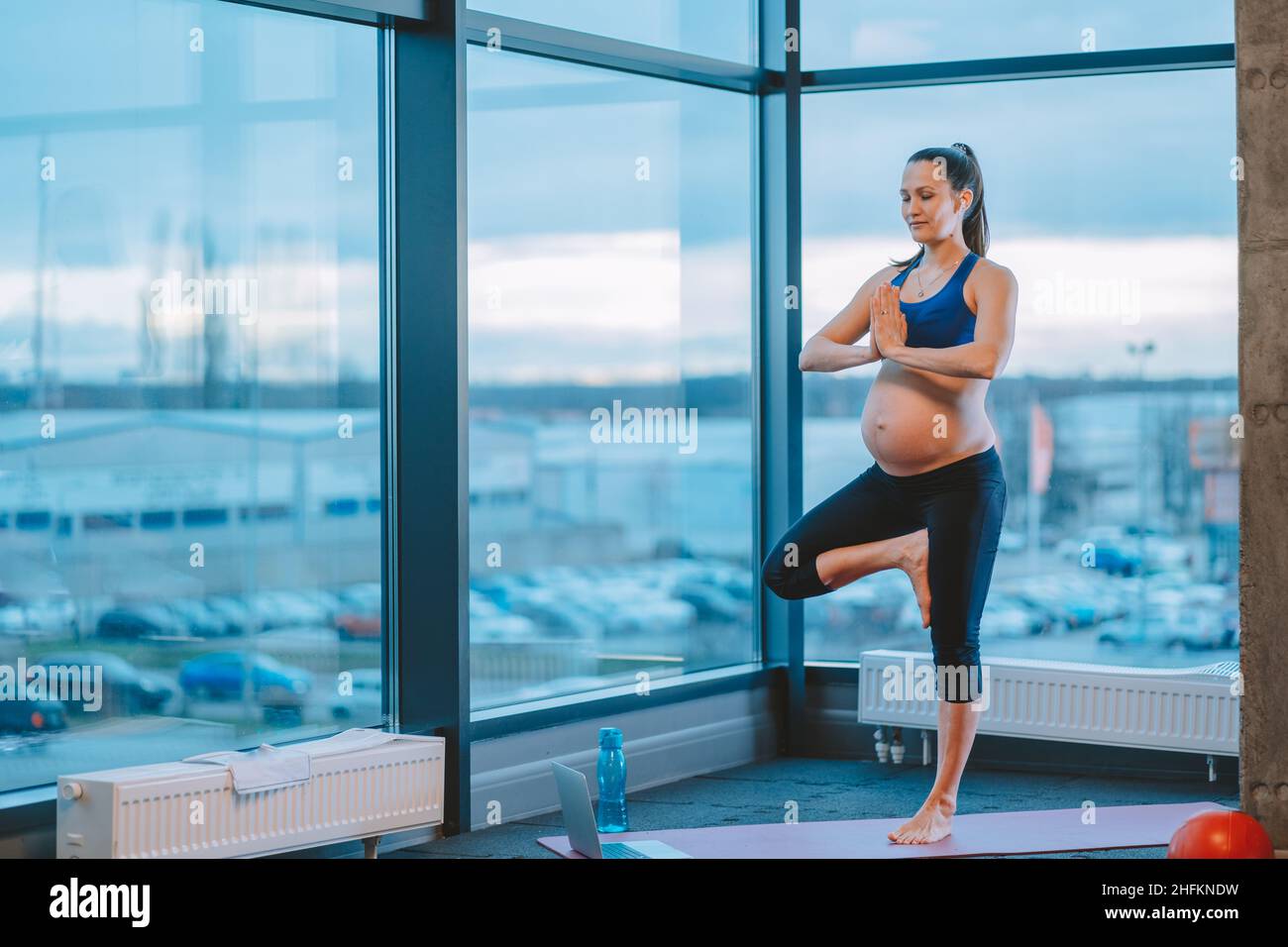 Mujer embarazada con las manos en namaste gesto de pie en la postura de árbol practicando yoga Foto de stock