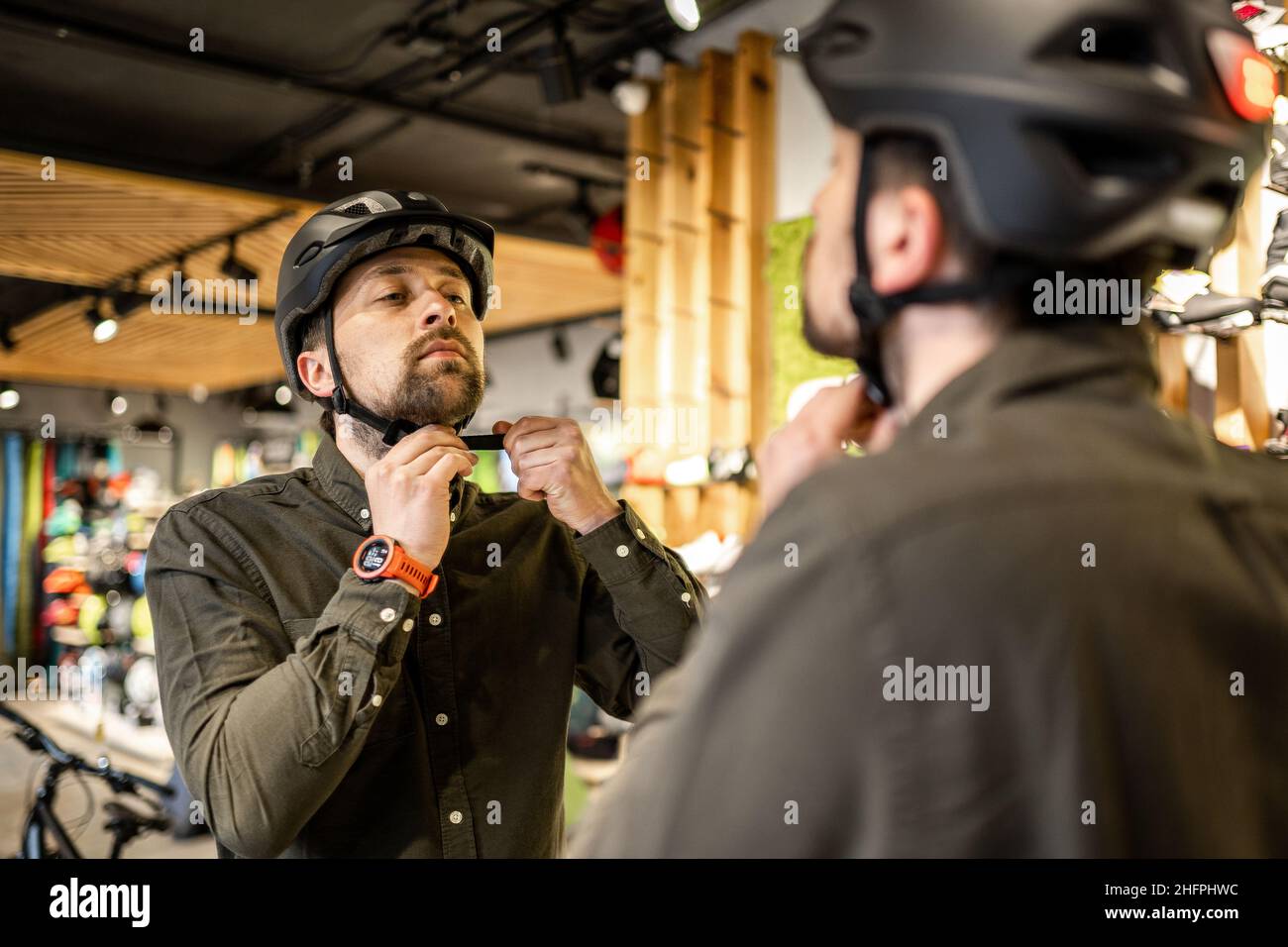 Hombre examinando cascos de bicicleta en una tienda de deportes mirándose  en espejo. Prueba de nuevo casco deportivo en la tienda de bicicletas.  Hombre poniendo casco de bicicleta en la tienda Fotografía