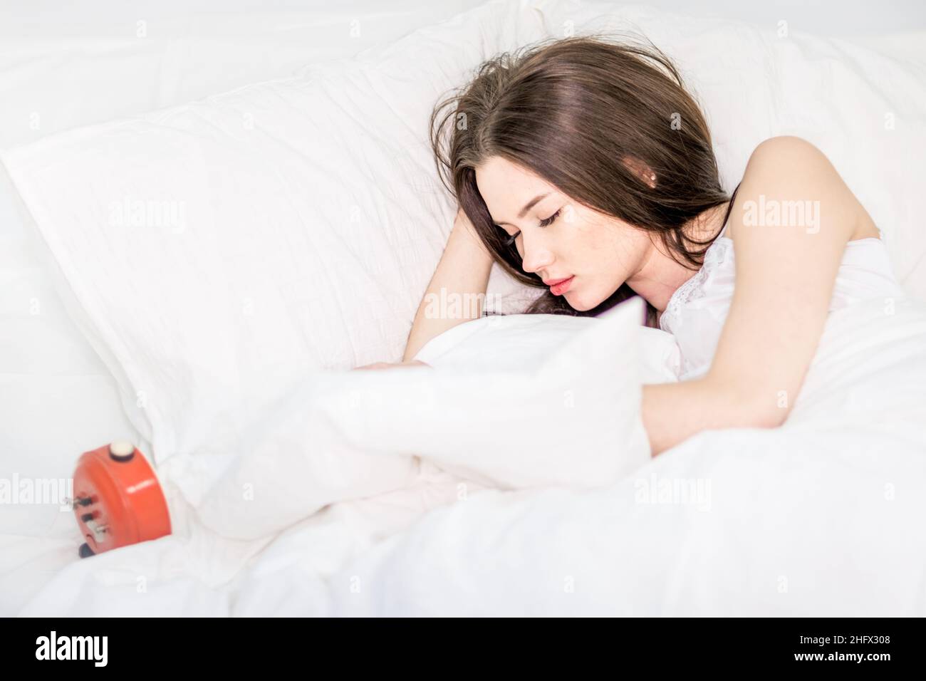 La morena duerme en una cama blanca. Una chica linda en pijama miente en la cama con sus ojos cerrados, un reloj despertador miente cerca. Buenos días. Relajación. Foto de stock