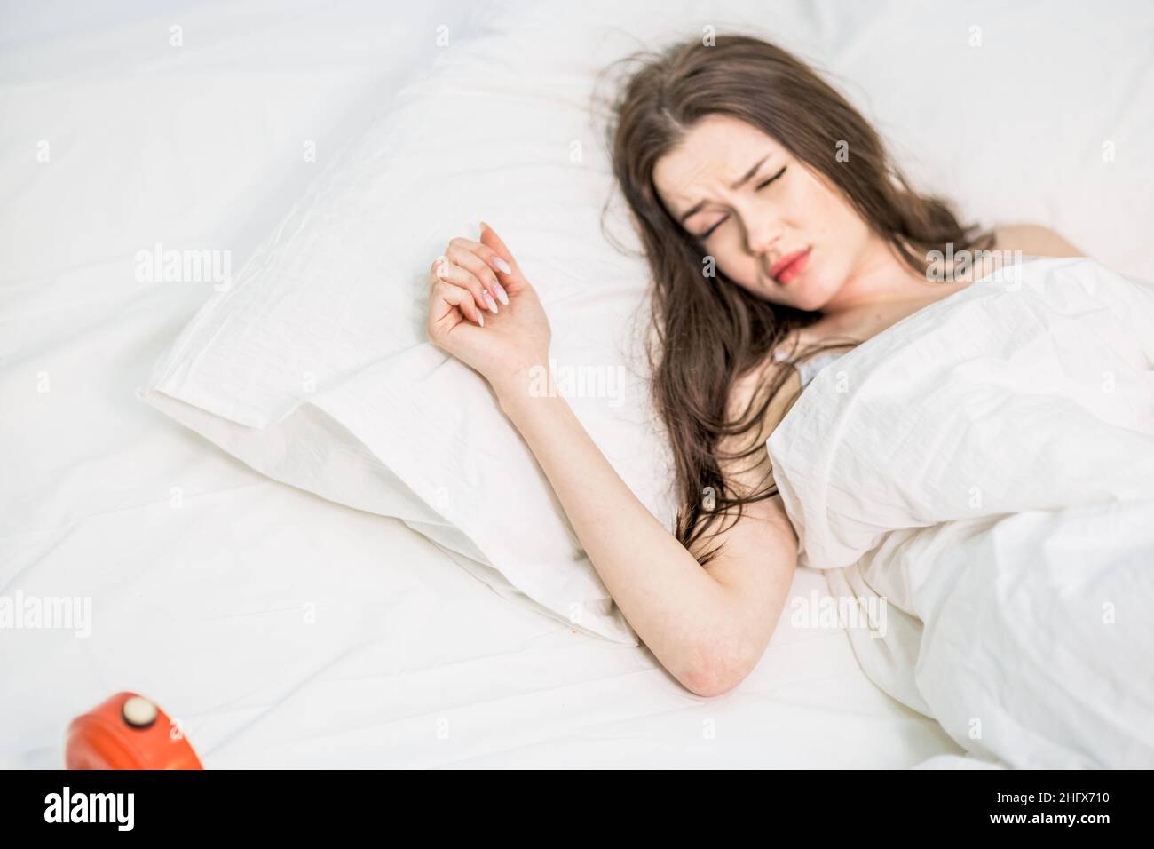 La mujer está molesta por el sonido fuerte. Buenos días. La morena se encuentra, se despierta del sonido del reloj despertador. Ruido fuerte. Cama blanca Foto de stock