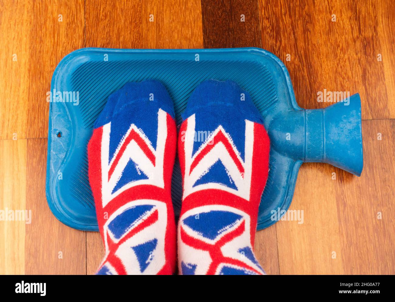 Persona que usa calcetines de Union Jack manteniendo los pies calientes en la botella de agua caliente. Aumento de energía, electricidad, precios de gas, costo de vida... concepto. Foto de stock
