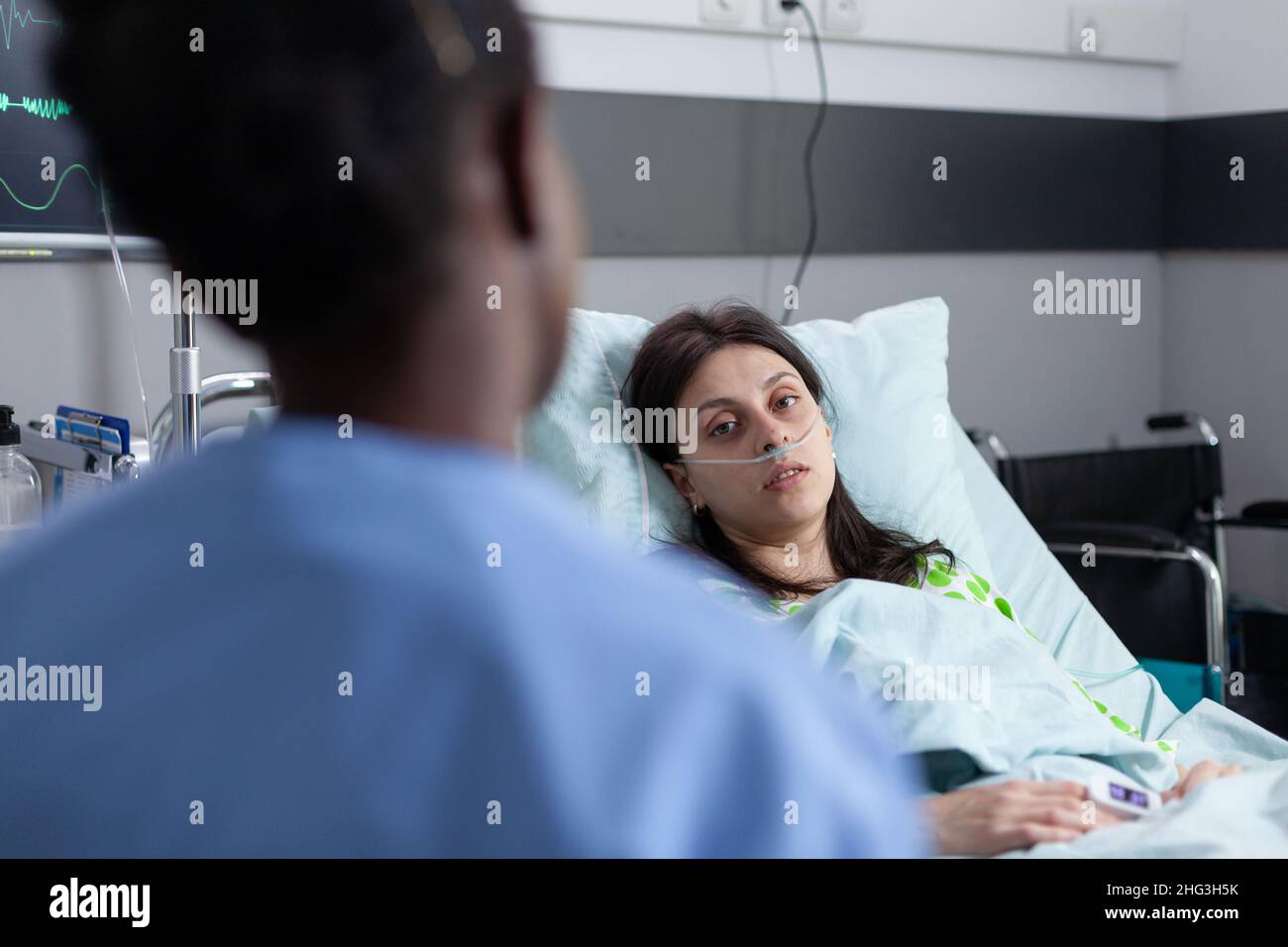 Vista sobre el hombro del paciente con cánula nasal que recibe oxígeno en la cama del hospital mirando a la enfermera que proporciona atención médica. Mujer con baja saturación de SpO2 en sala de hospital privada. Foto de stock