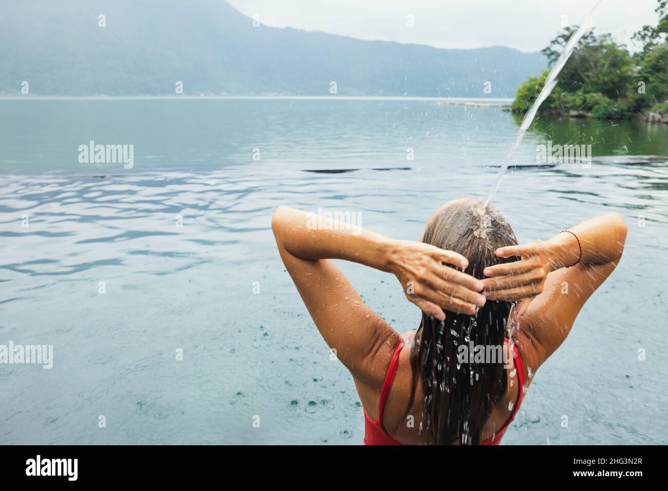 Una mujer joven se relaja bajo el agua corriente en la piscina infinita con hermosas vistas al lago. Balneario natural de aguas termales junto al volcán Batur. Tour turístico en Kintaman Foto de stock