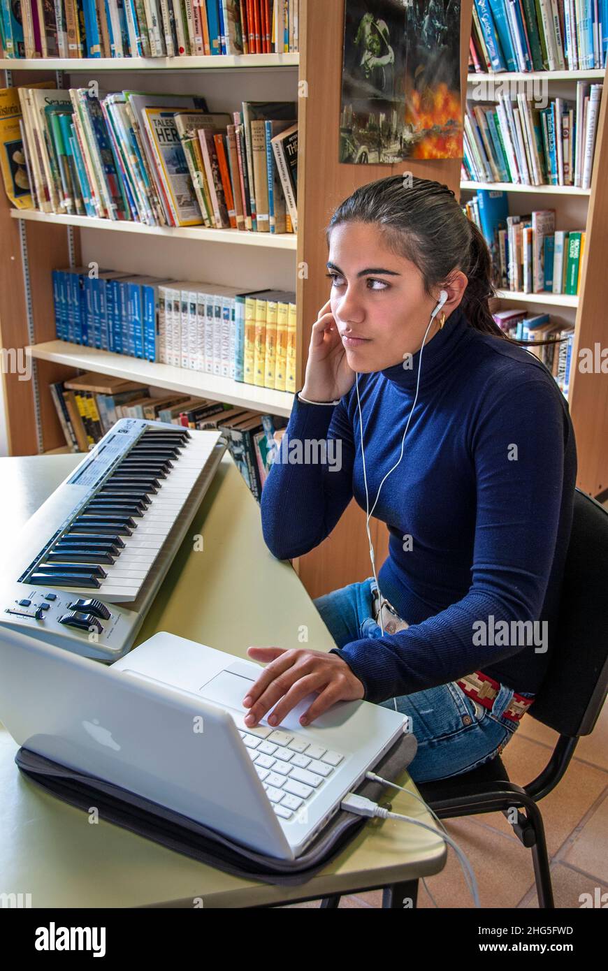 Estudiante de escuela adolescente hispana con auriculares escuchando y componiendo con computadora portátil y teclado de música electrónica en la biblioteca de la escuela Foto de stock
