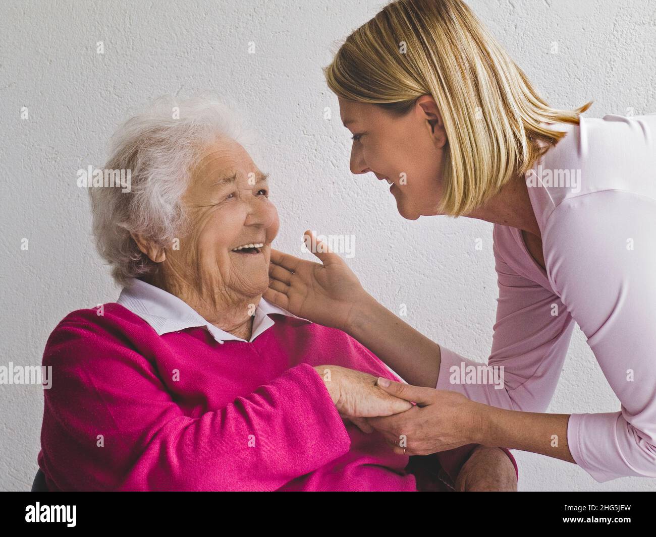 ANCIANA SEÑORA con joven cuidador cuidador visitante mujer cariñosamente táctil tocar las manos mejilla saludo encantador feliz sonrisa anciana anciana anciana anciana anciana jubilada en luz natural Foto de stock