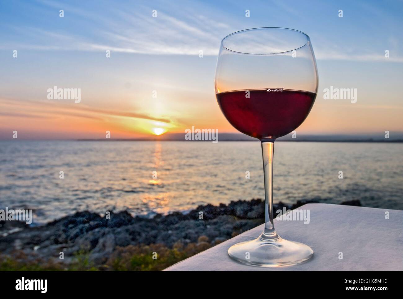 Copa de vino tinto en la mesa del restaurante al atardecer con vistas al paisaje marino detrás Foto de stock