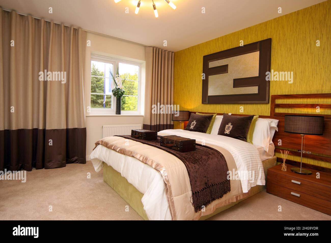 Fondo de pared amarillo dorado en un dormitorio, estilo de vida, cojines, cama doble king size, luces de cabecera, espejo de pared, flores en la ventana. Foto de stock