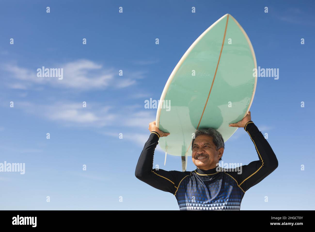 Vista en ángulo bajo del hombre mayor biracial llevando tablas de surf sobre la cabeza en la soleada playa contra el cielo azul Foto de stock