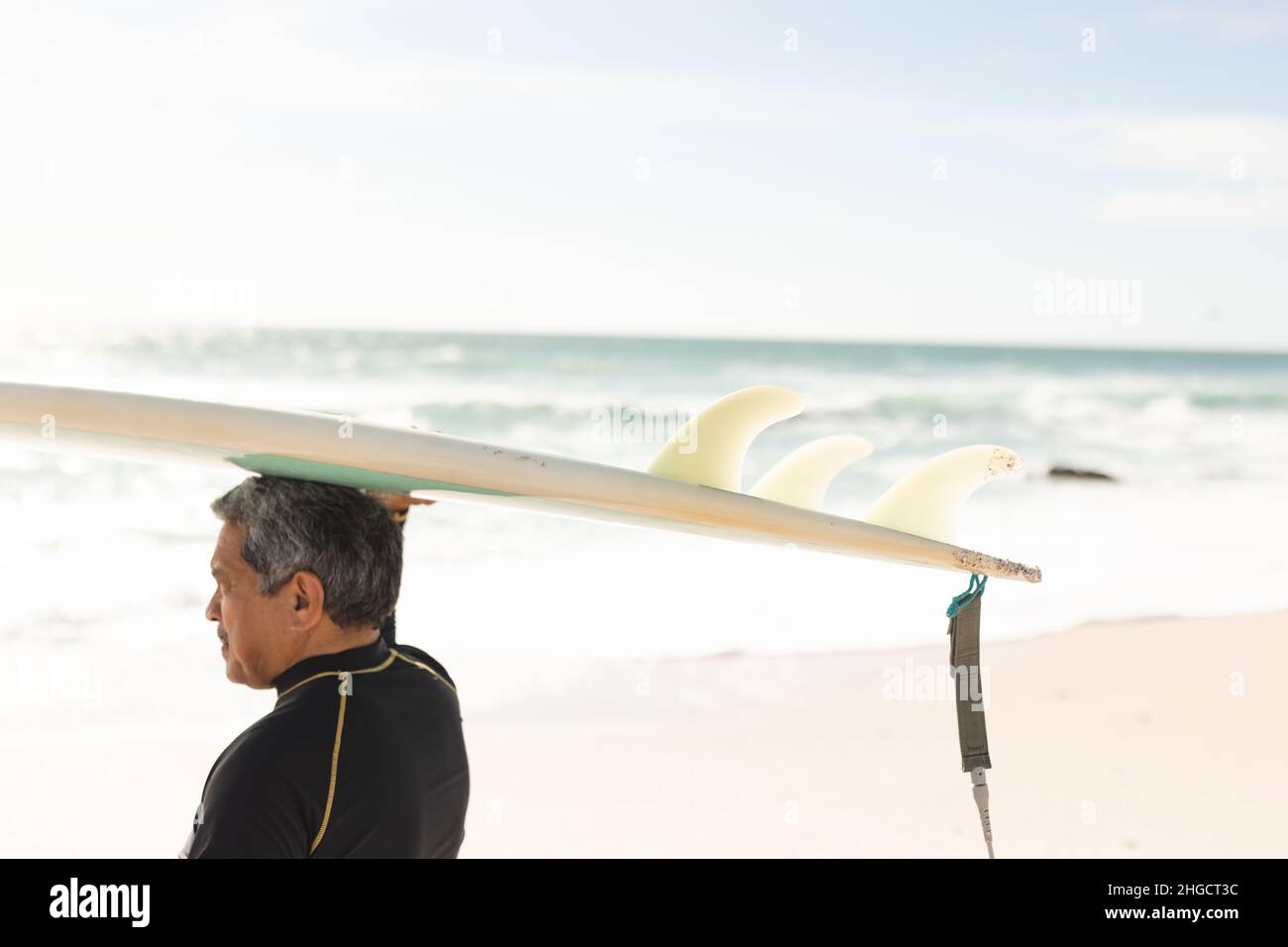 Hombre de edad biracial cargando tabla de surf sobre la cabeza en la playa contra el cielo durante el día soleado Foto de stock