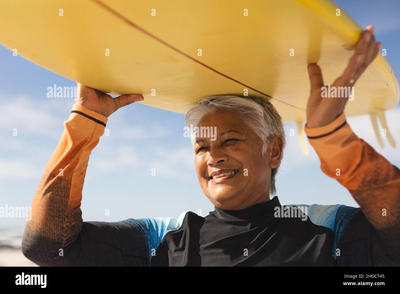 Feliz mujer de edad biracial cargando tabla de surf amarilla sobre la cabeza en la playa durante el día soleado Foto de stock
