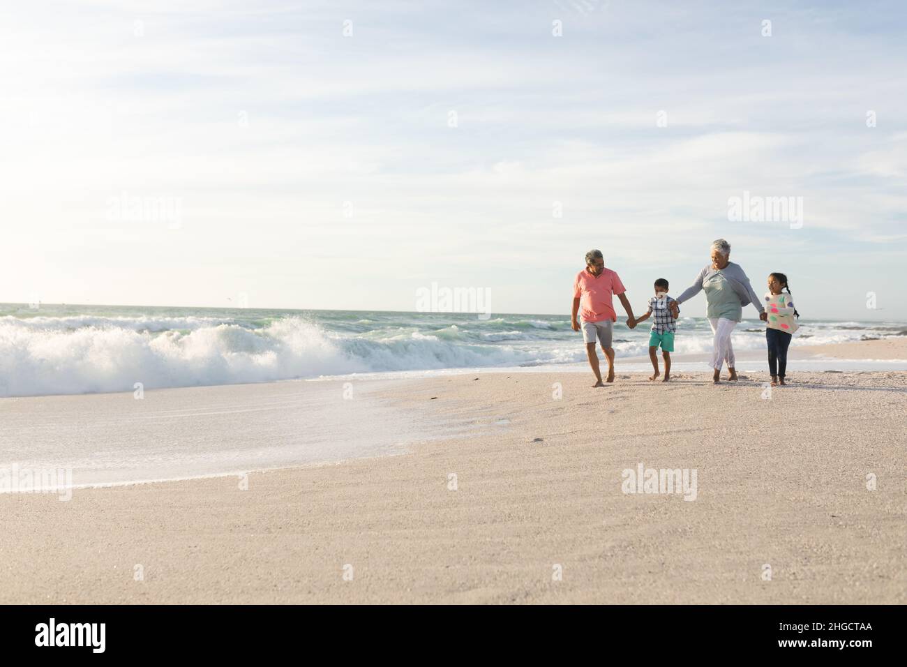 Abuelos multirraciales sosteniendo manos con nietos caminando en la playa contra el cielo Foto de stock