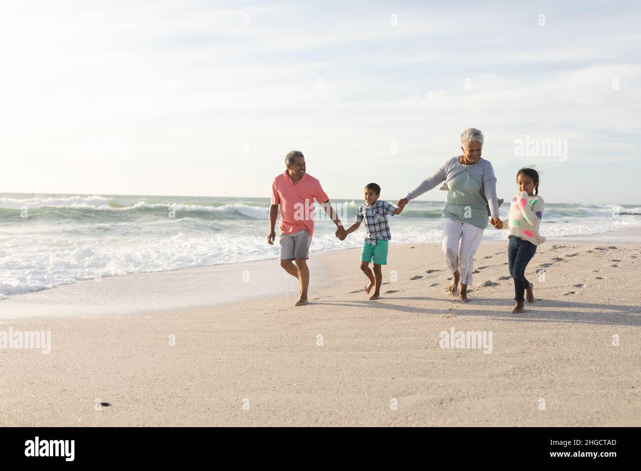 Nietos multirraciales que sostienen las manos con abuelos que caminan en la playa contra el cielo Foto de stock