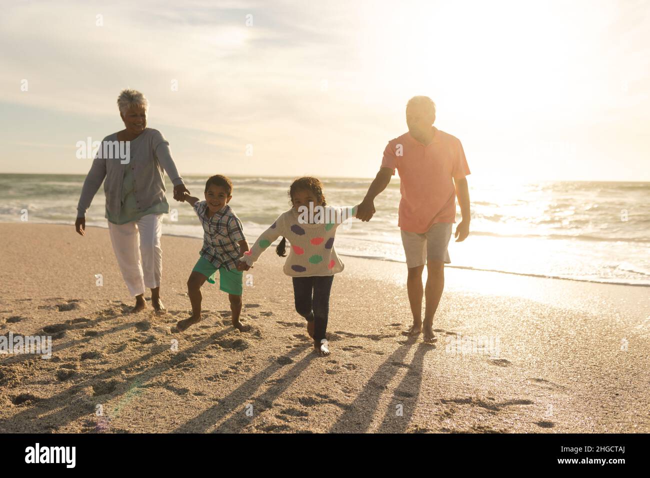 Nietos multirraciales sosteniendo las manos con abuelos caminando en la playa durante la puesta del sol Foto de stock