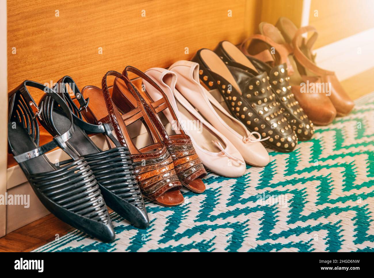 Varios zapatos para mujer con diferentes diseños colocados en fila sobre alfombra de colores en la pared de madera en la habitación de luz en casa Foto de stock