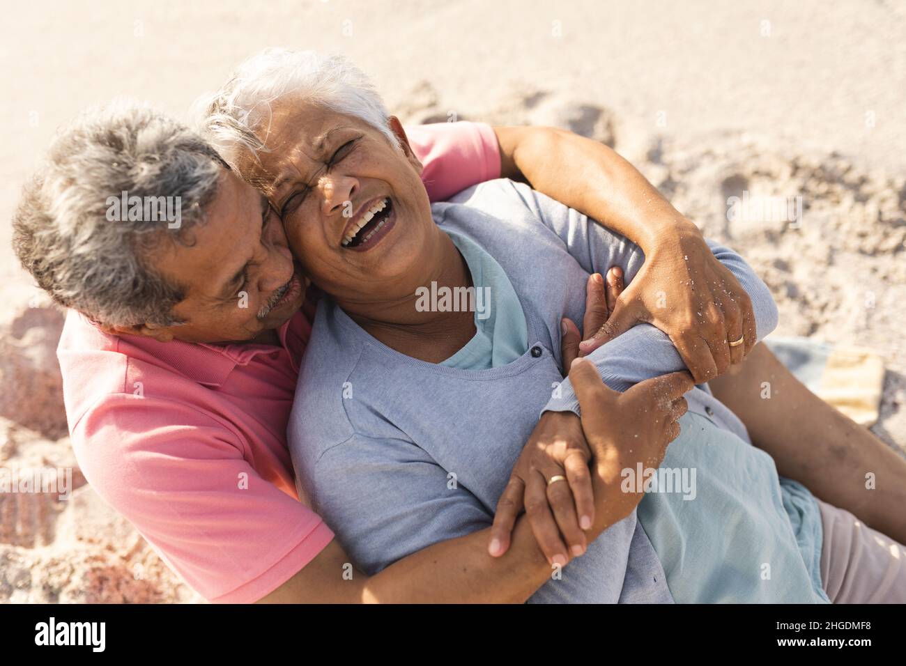 Hombre mayor biracial abrazando a una mujer alegre de detrás mientras se sentaba en la playa en un día soleado Foto de stock