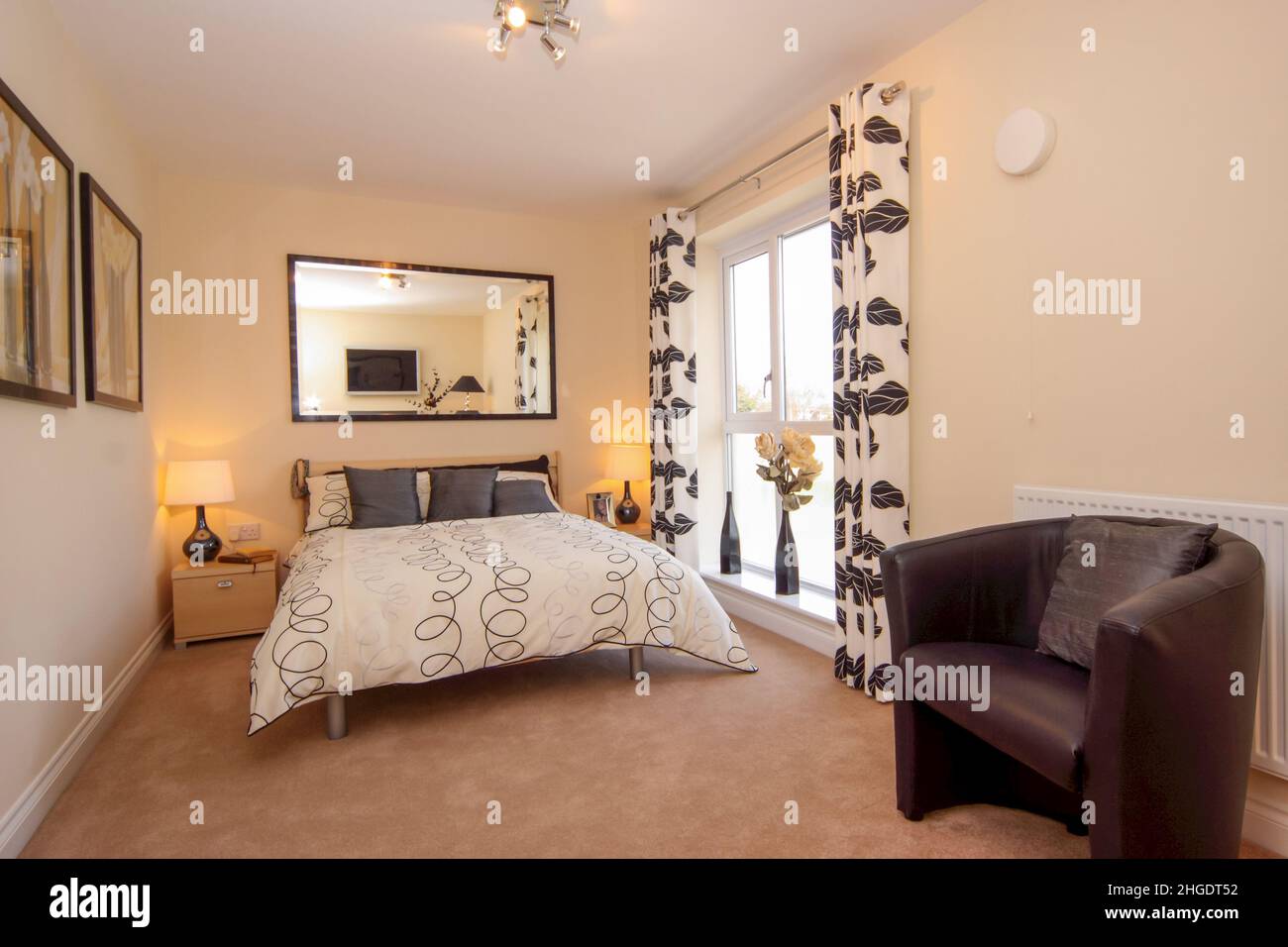 Dormitorio en tonos neutros, cama doble king size, bajo alféizar, cortinas con diseño de hojas, espejo de pared, alfombra beige. Foto de stock